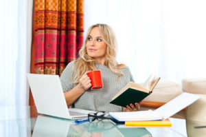 attractive woman with cup of coffee, book and laptop sitting at her home desk and working