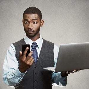 Closeup portrait shocked, surprised business man reading bad news on smart, mobile, cell phone holding laptop computer isolated black grey background. Human face expression emotion corporate executive