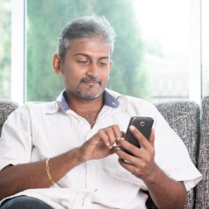 Smartphone social media concept. Indian mature guy using mobile phone. Asian man relaxed and sitting on sofa indoor.